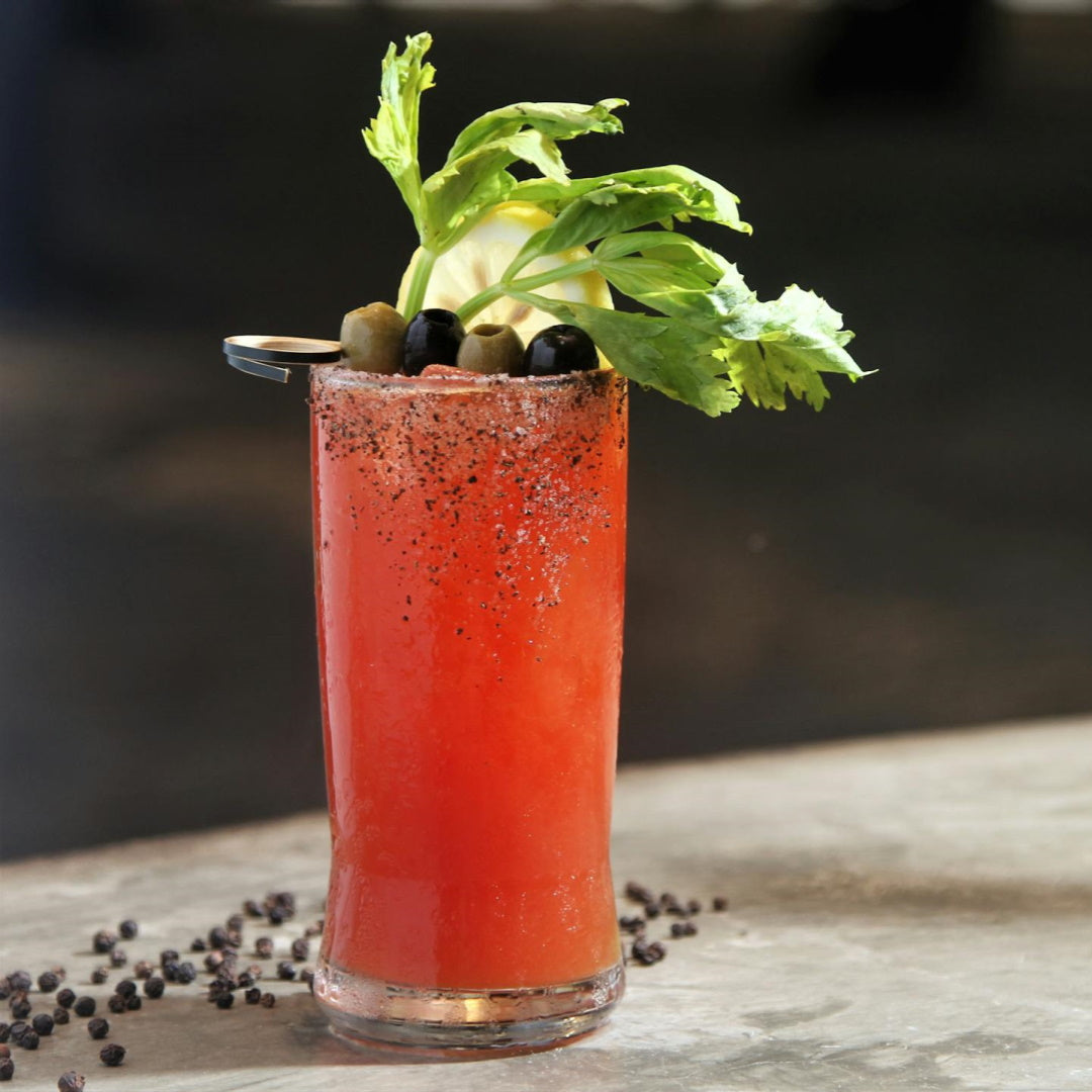 Tall Bloody Mary with cracked-pepper rim, celery stalk, lemon wheel and olives on a skewer; peppercorns scattered on table.