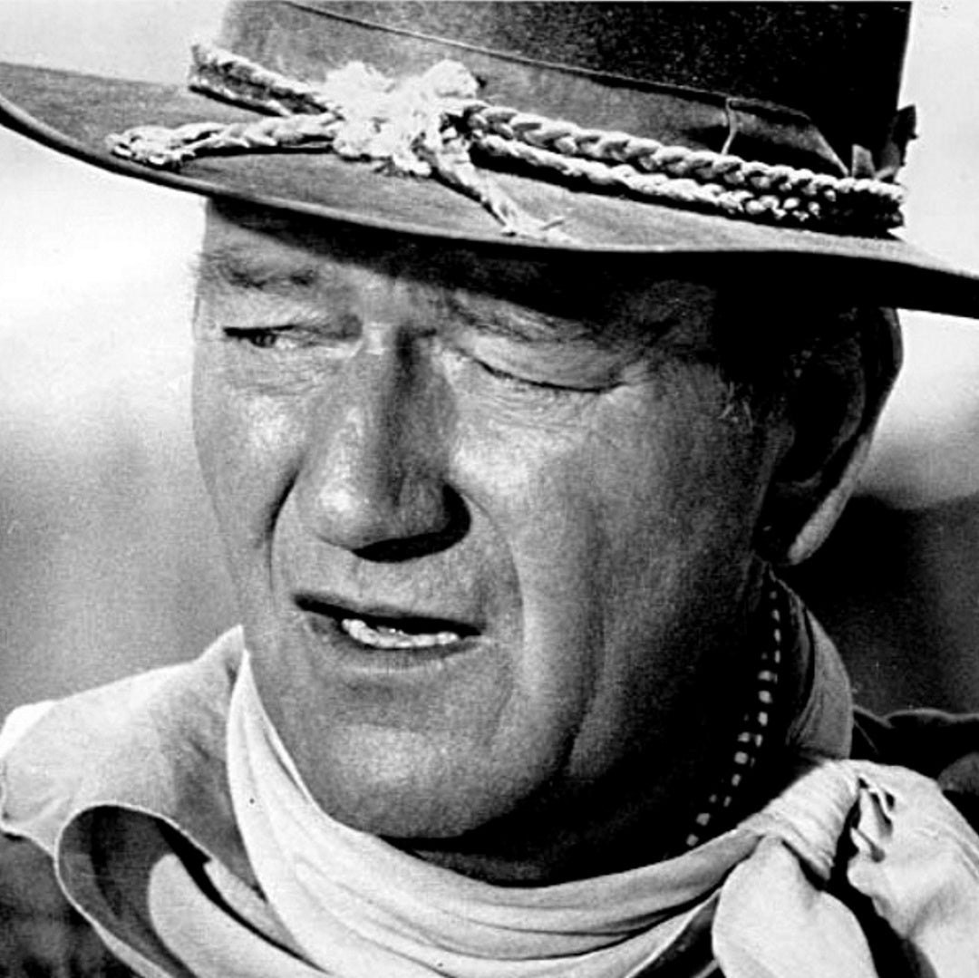 Close-up of John Wayne wearing a cowboy hat with braided band and a neckerchief, squinting and looking left.