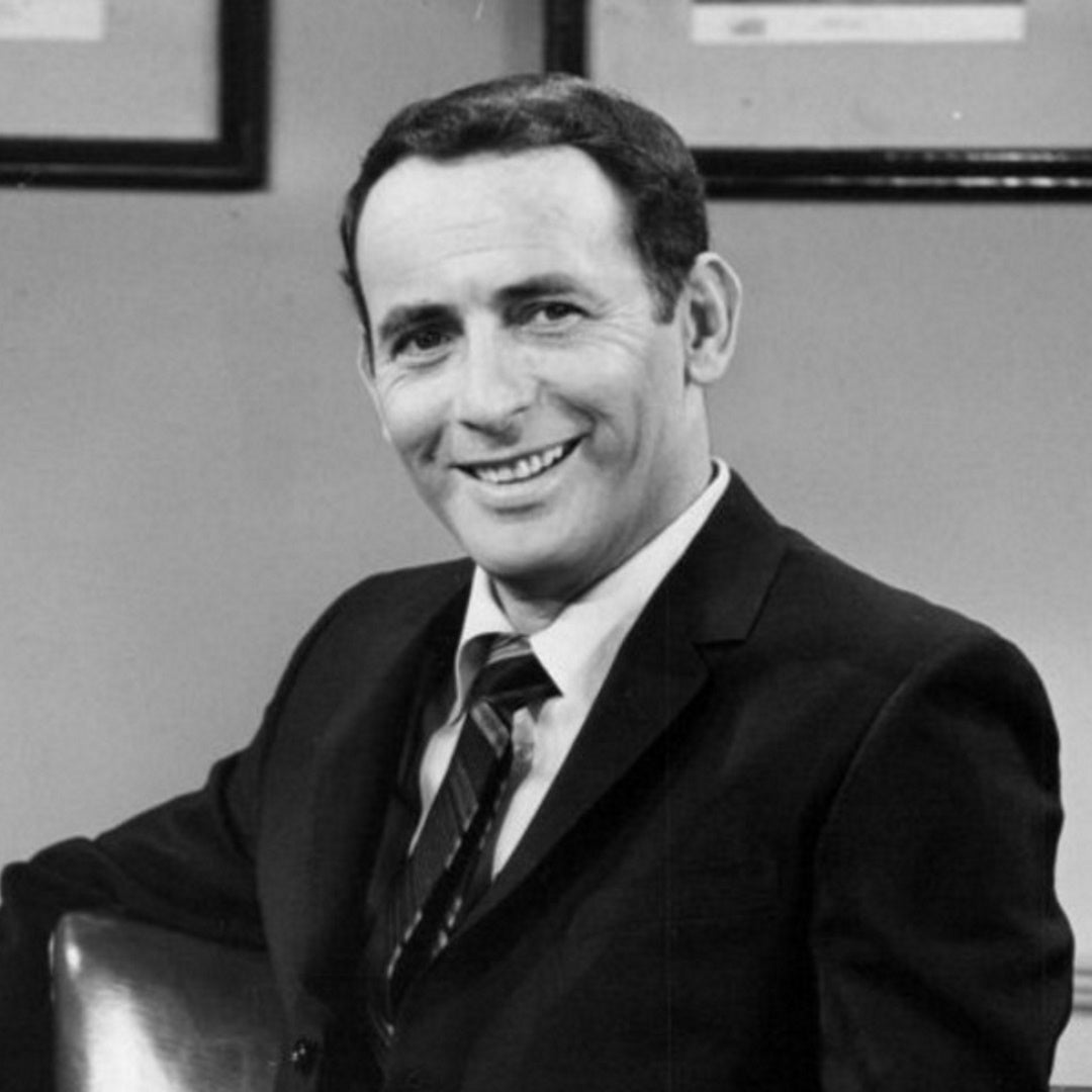 Black-and-white portrait of Joey Bishop in a dark suit, seated three-quarter view in an office with framed certificates behind.