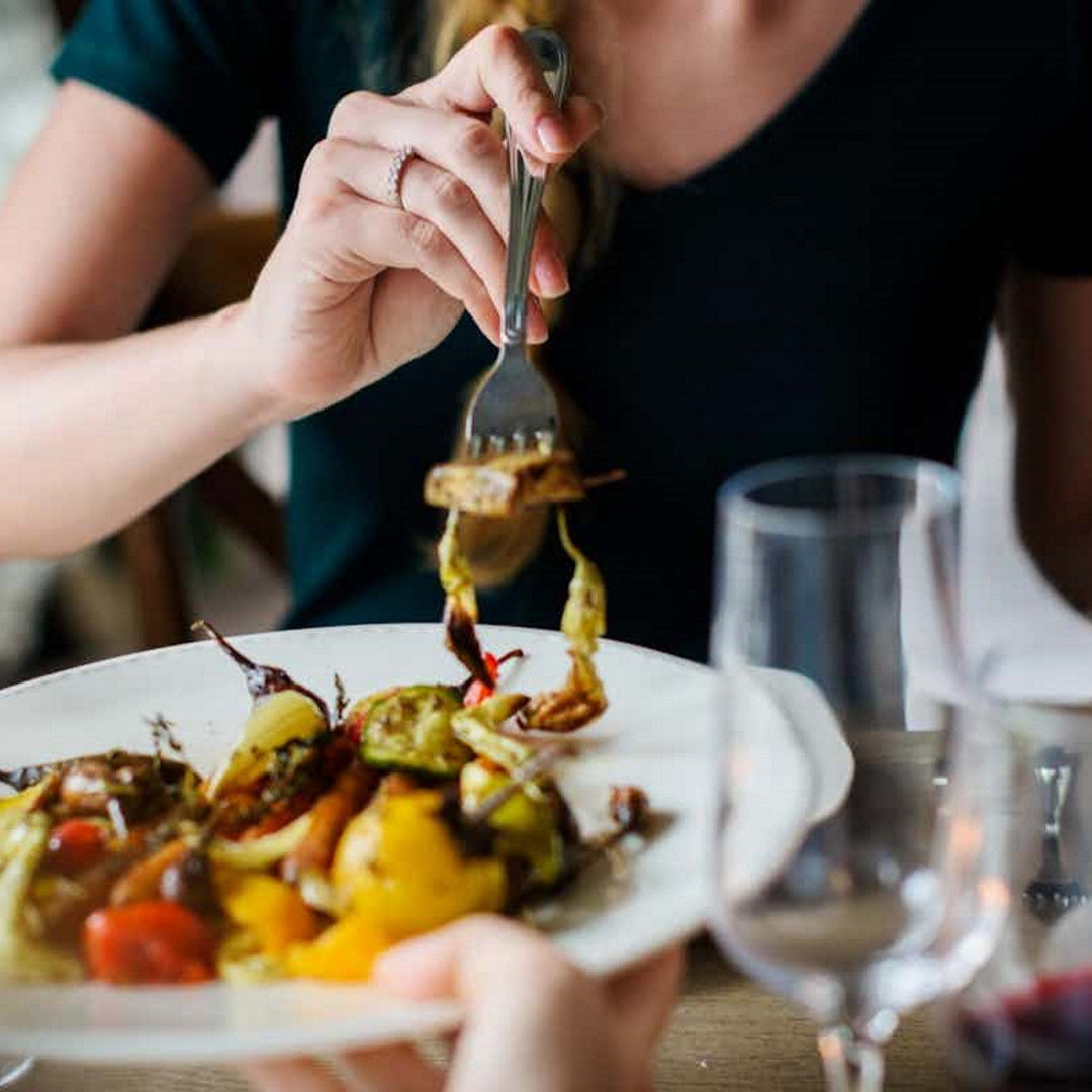 Hand with fork lifting roasted vegetables from a white plate; wine glasses and another hand visible.