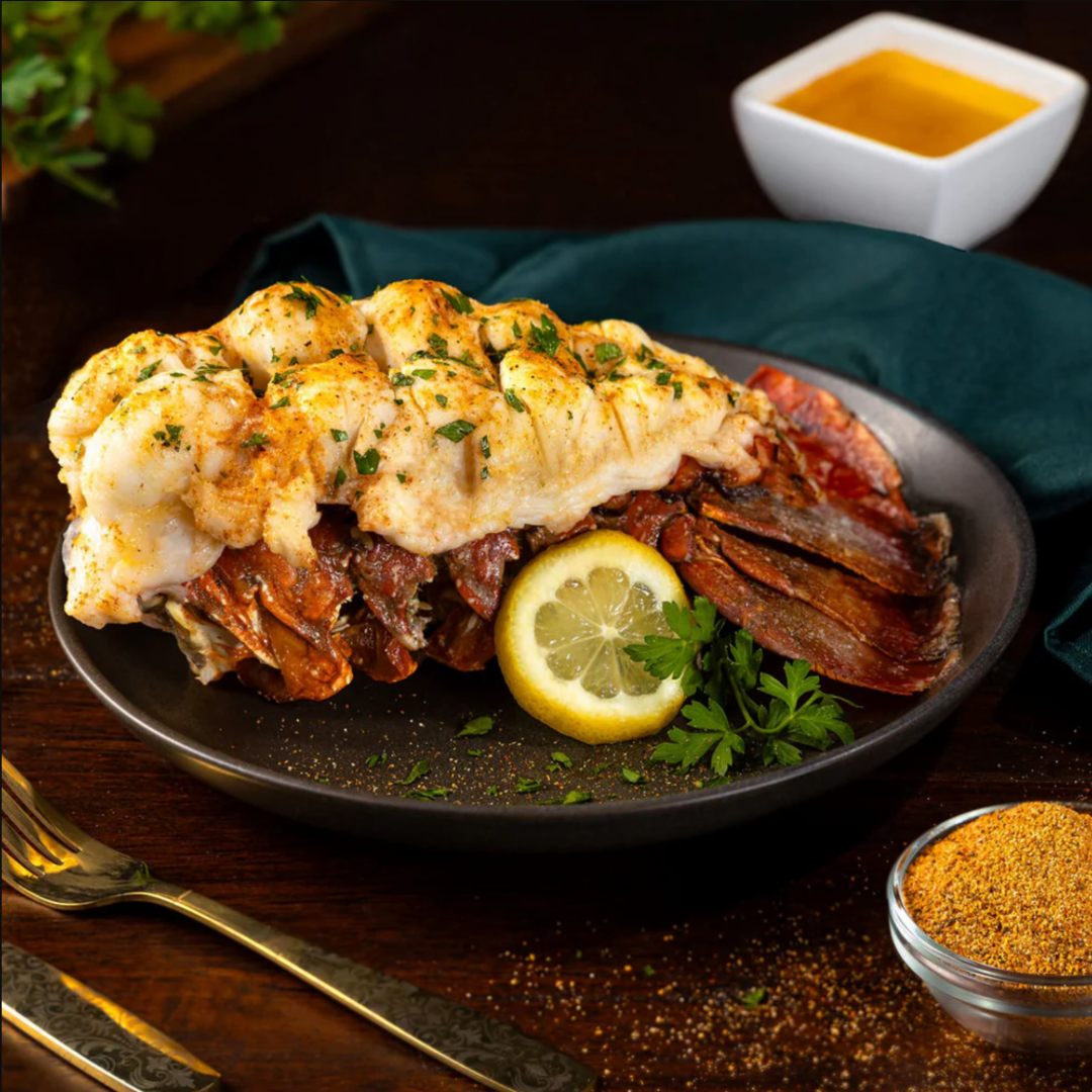 Grilled lobster tail served on its shell with herbs, lemon slice, butter sauce and a small bowl of seasoning on a black plate