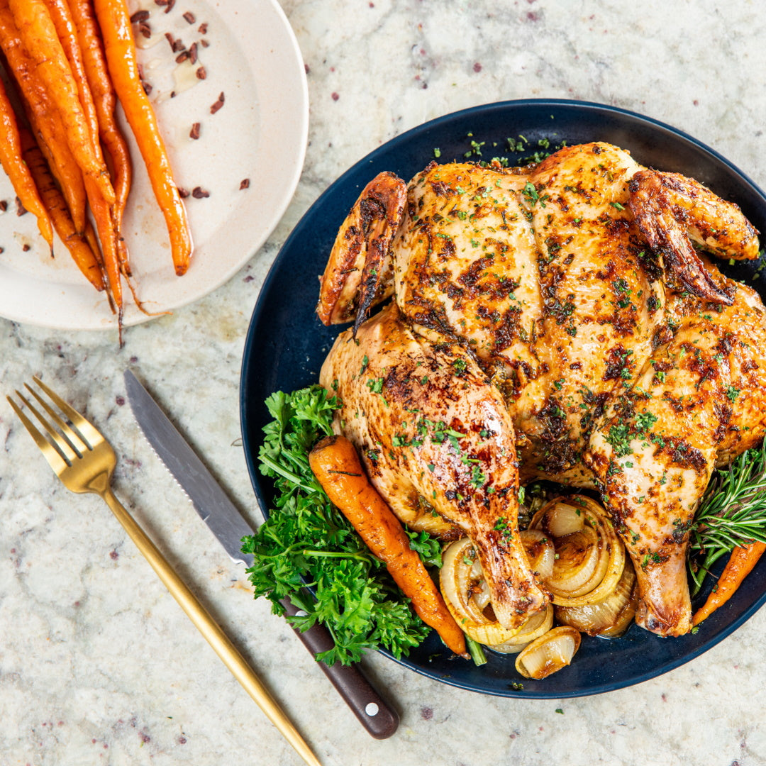Whole roasted chicken on a dark plate with roasted carrots, caramelized onions, parsley and rosemary