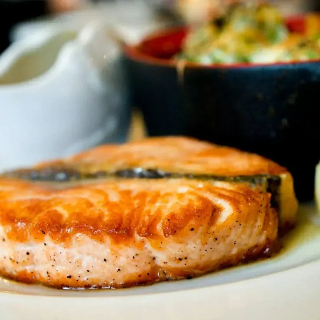 Pan-seared salmon steak on a plate with a blurred sauce boat and side salad bowl in background.