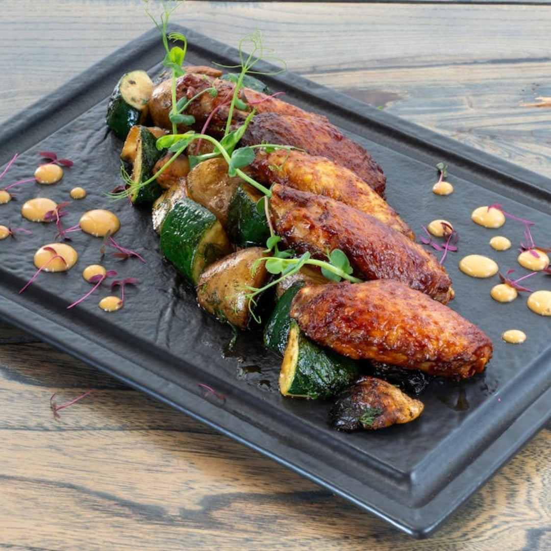 Grilled chicken wings with roasted zucchini, potatoes, microgreens and mustard dollops on a black rectangular platter