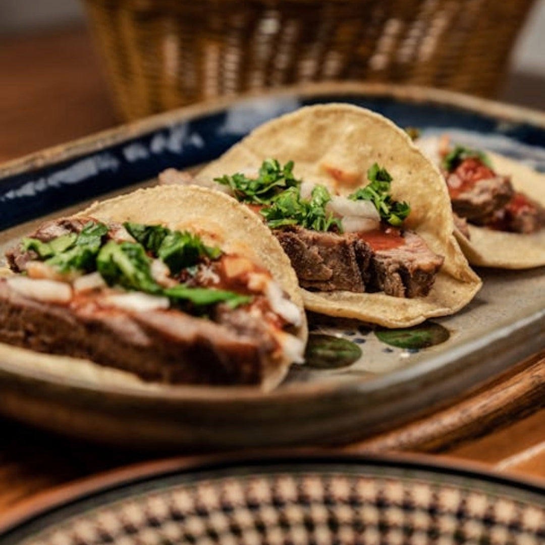 Three carne asada tacos on corn tortillas topped with chopped onion and cilantro on a ceramic plate