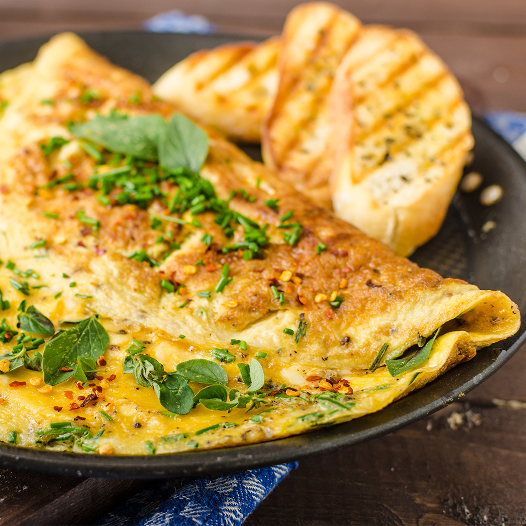 Folded omelette topped with fresh herbs and crushed red pepper, served with grilled garlic bread on a skillet.