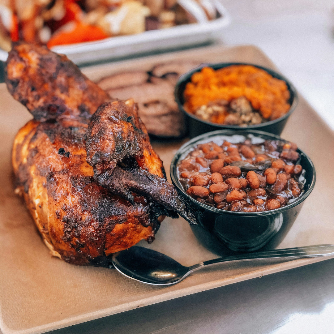 Smoked half chicken on a tray with a bowl of BBQ baked beans and a spoon; sweet potato mash and brisket behind.