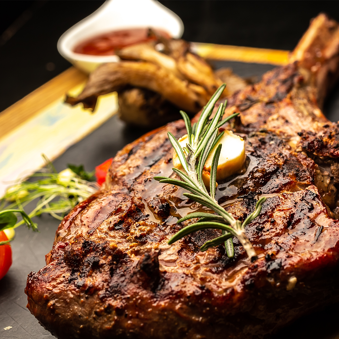 Grilled bone-in ribeye with herb crust, roasted garlic, rosemary sprig and mushrooms on a serving board