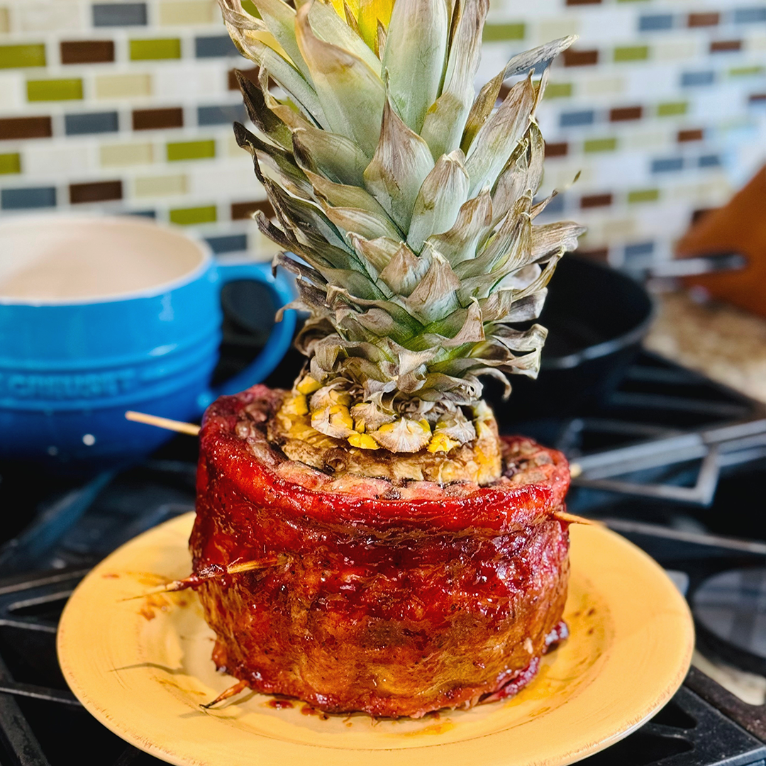 Smoked rib roast wrapped in bacon, glazed and topped with a halved pineapple crown on a yellow plate