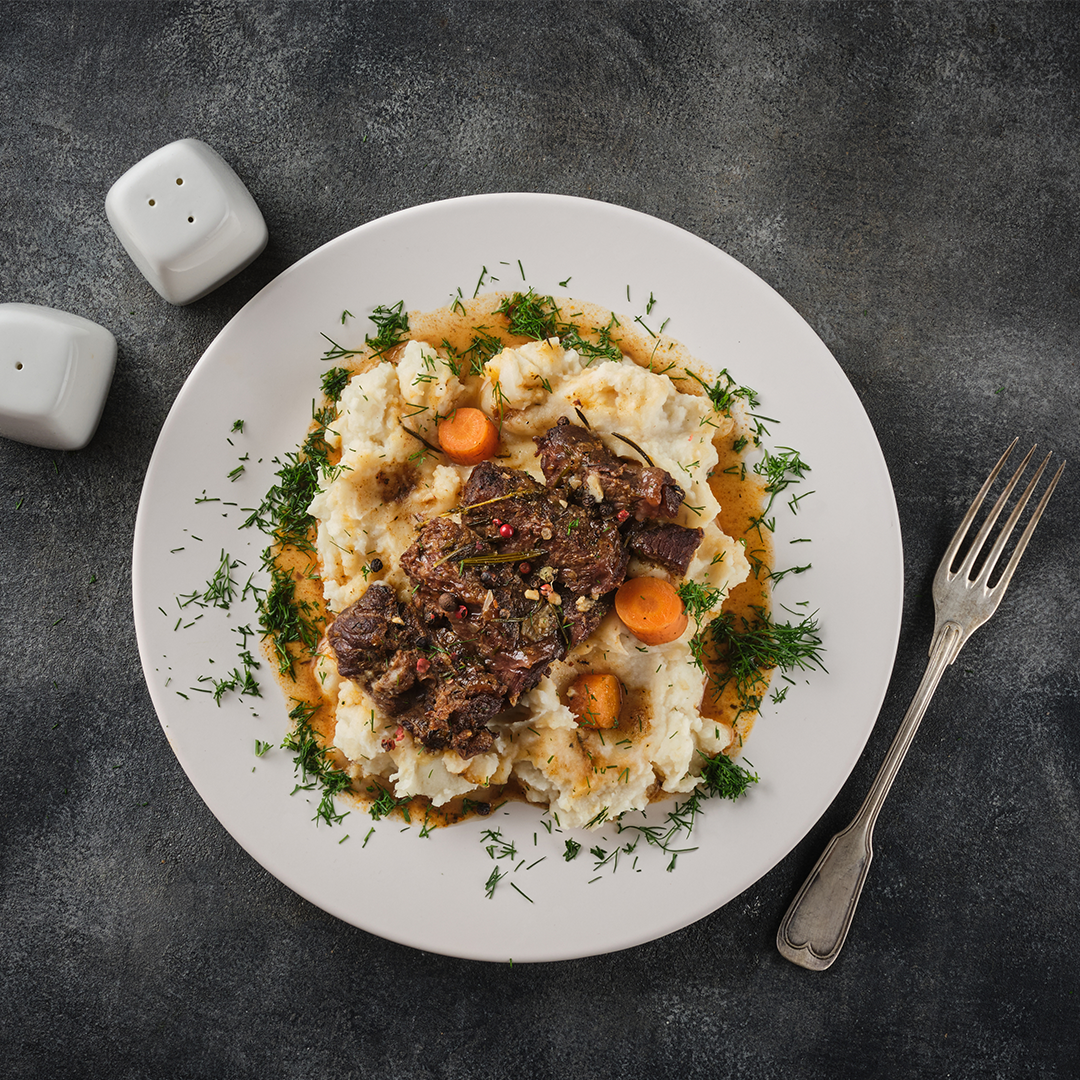 Braised pot roast on mashed potatoes with carrots, gravy and chopped dill, with salt shakers and a fork on a dark tabletop.