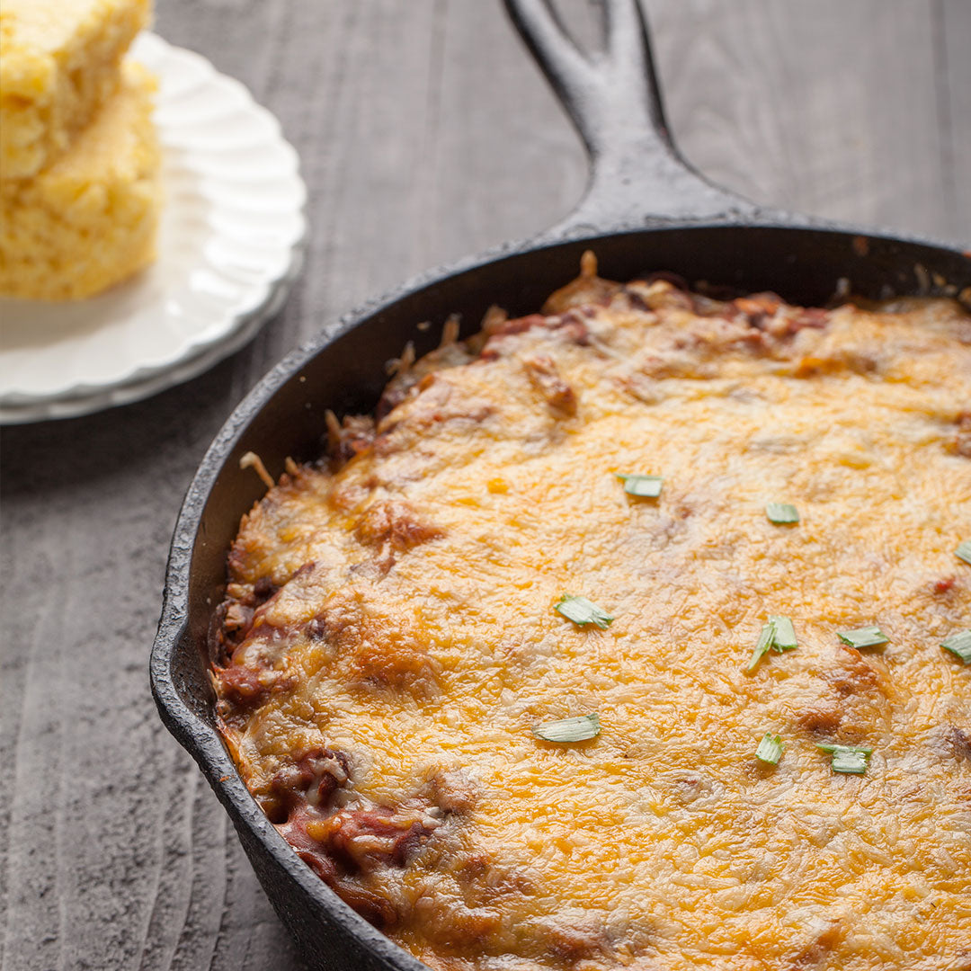 Cheesy cornbread chili bubbling in a cast iron skillet with a slice of cornbread on a plate beside.