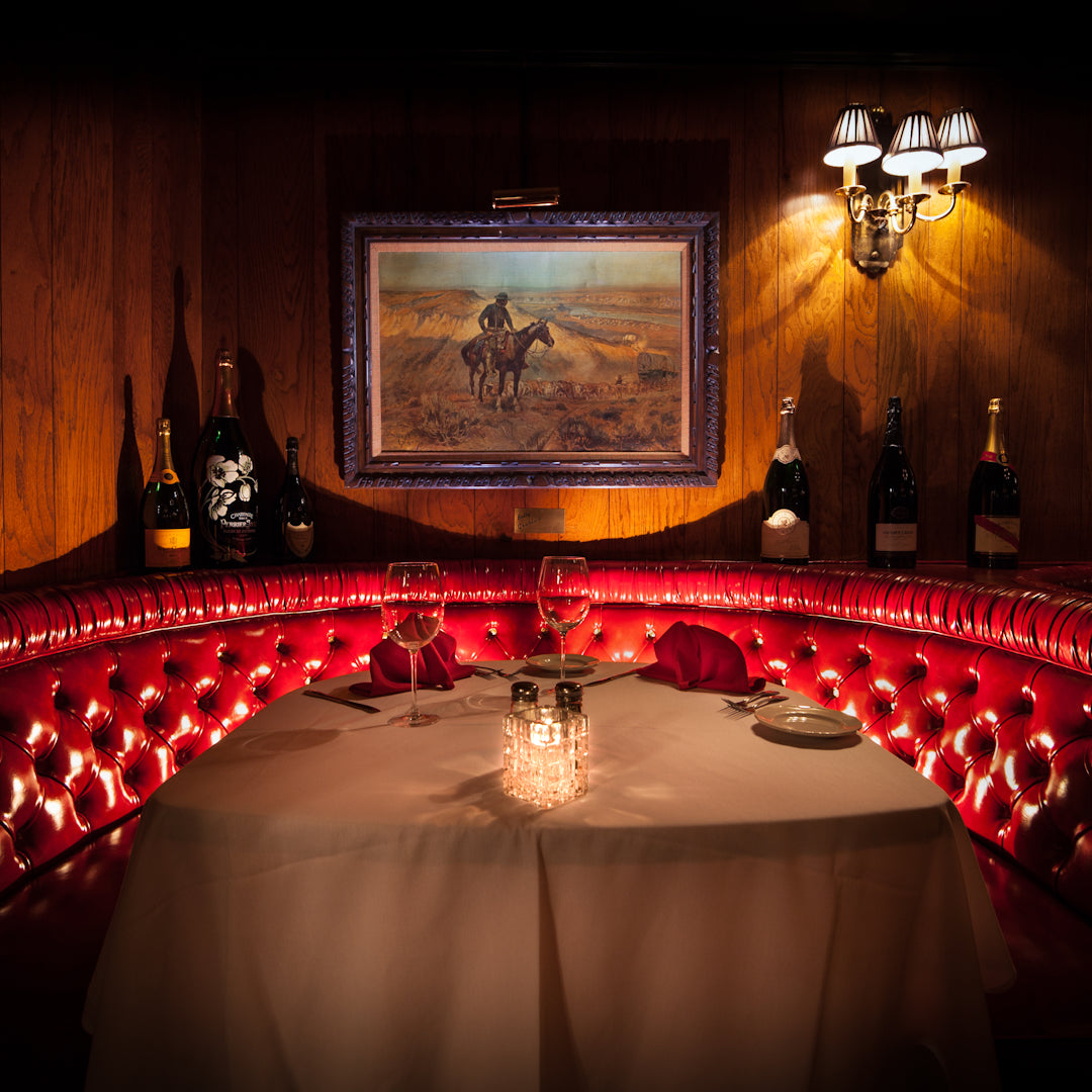 Red tufted leather booth set for two with white tablecloth at Golden Steer Las Vegas, wine glasses, folded red napkins and a candlelit centerpiece in a wood-paneled dining nook